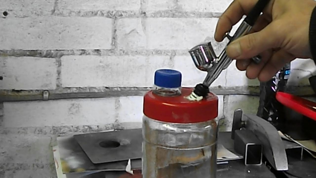 A close-up of an airbrush being cleaned at a hobby desk with cleaning cups, cotton swabs, and a small bottle of airbrush cleaner nearby