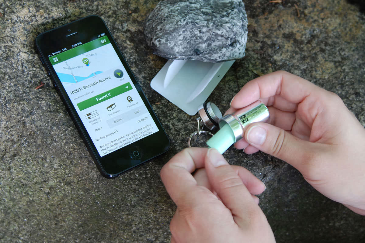A person holding a smartphone with a geocaching map open while standing at the edge of a wooded trail, ready to start searching