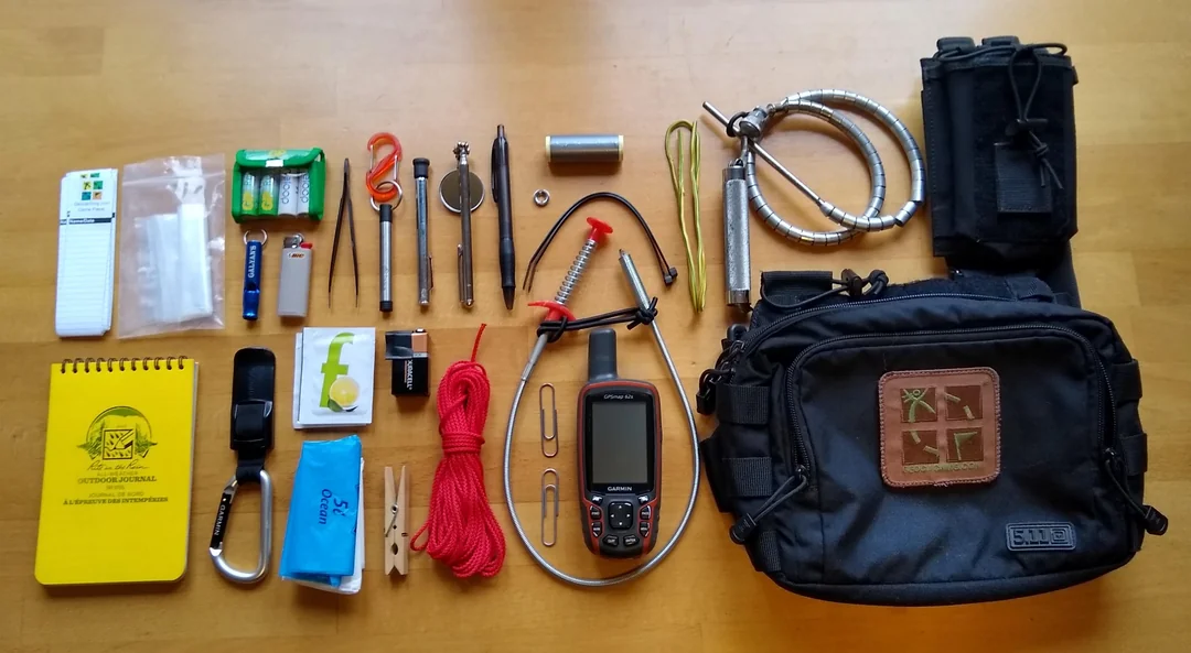 A small pouch on a picnic table holding a pen, a tiny flashlight, tweezers, and hand sanitizer next to a smartphone
