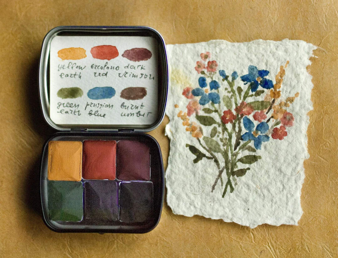 A small travel paint palette open on a park bench next to a sketchbook with a simple line drawing of nearby buildings