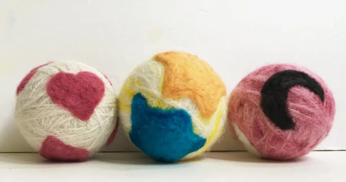 Small needle felted wool ball resting on a foam mat next to a felting needle handle, showing a firm round shape with slight fuzz