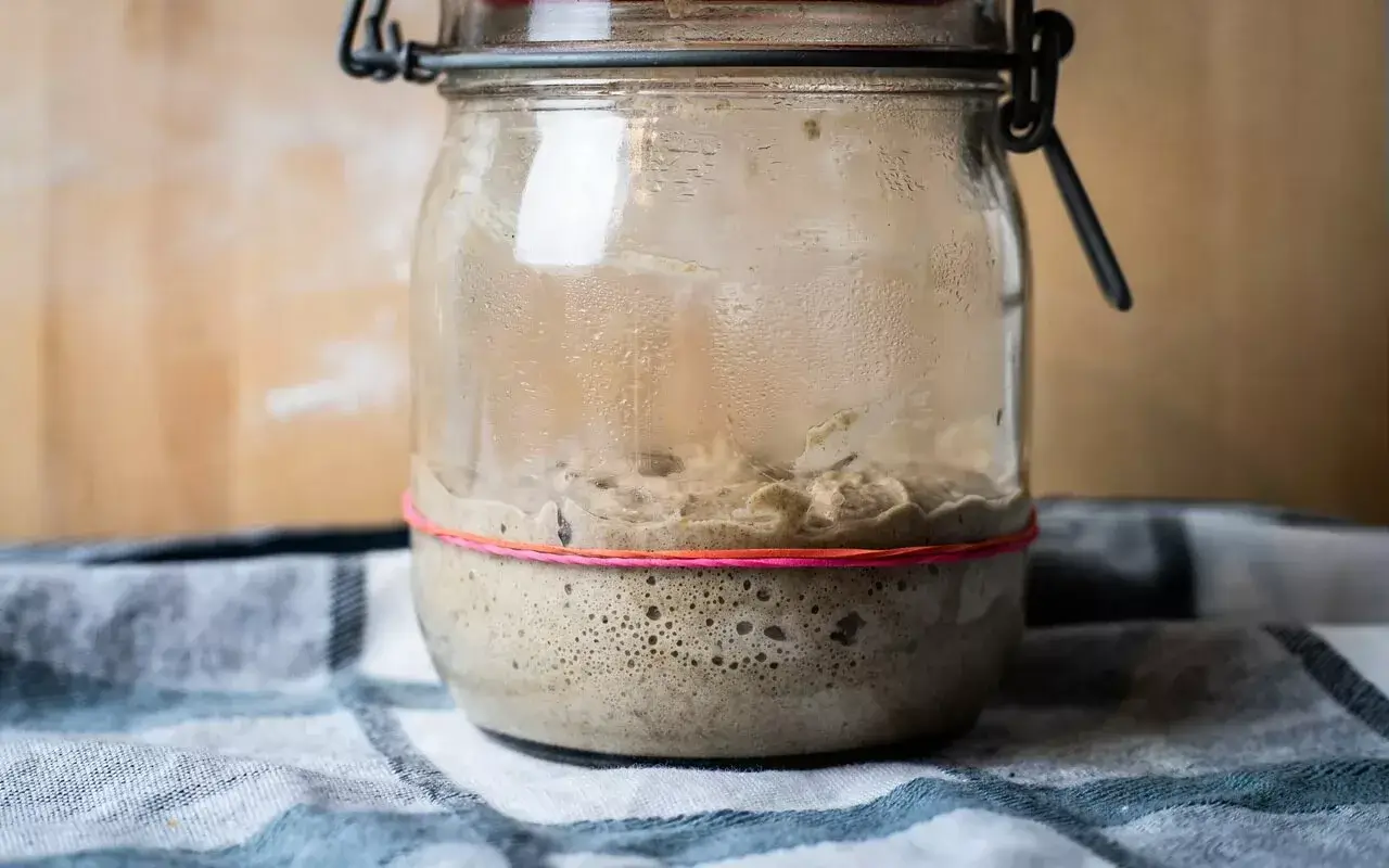 Someone feeding a sourdough starter on a kitchen scale, pouring flour into a jar next to a small bowl of water