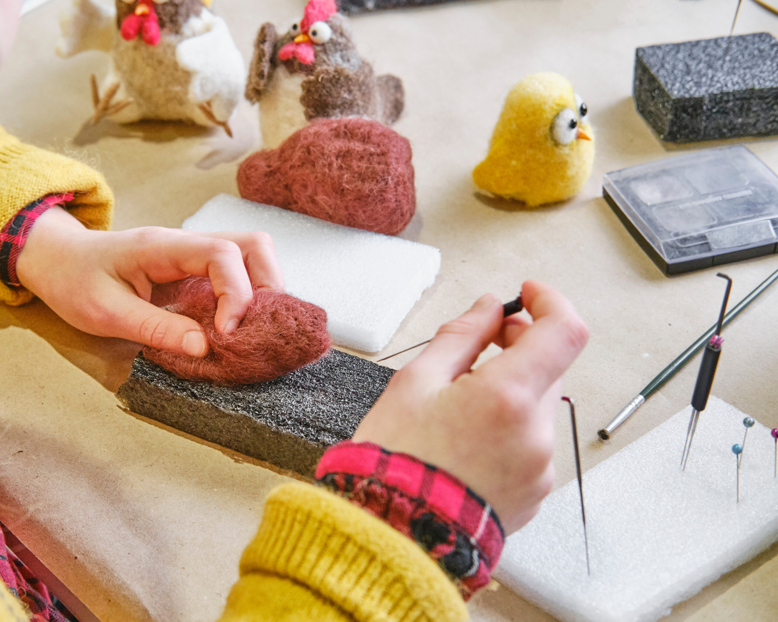 Tidy needle felting workspace on a dining table with a desk lamp, foam mat, wool bundles in a small tray, and needles stored in a cork